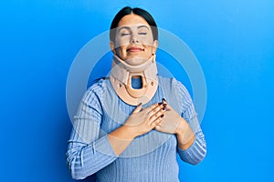 Beautiful brunette woman wearing cervical collar smiling with hands on chest with closed eyes and grateful gesture on face