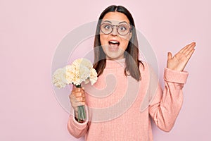 Beautiful brunette woman wearing glasses holding bouquet of flowers over pink background celebrating victory with happy smile and