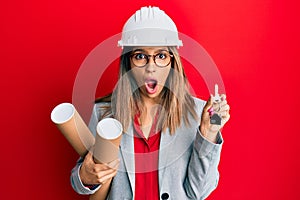 Beautiful brunette woman wearing safety helmet holding blueprints afraid and shocked with surprise and amazed expression, fear and