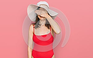 Beautiful brunette woman wearing swimsuit and summer hat smiling and laughing with hand on face covering eyes for surprise