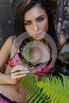Beautiful brunette woman with wet hair in red bathing suit lying in the jungle shadows on the ground with a branch of fern