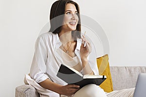 Beautiful brunette woman in white shirt sits on sofa in home office, holds pencil, black notepad in her hand.