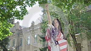 Beautiful brunette woman wrapped in American flag raising hand up. Portrait of confident charming lady showing liberty