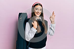 Beautiful brunette young musician woman wearing guitar case doing ok sign with fingers, smiling friendly gesturing excellent