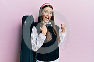 Beautiful brunette young musician woman wearing guitar case pointing thumb up to the side smiling happy with open mouth