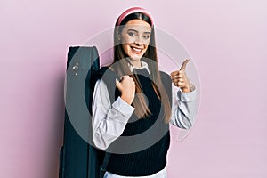 Beautiful brunette young musician woman wearing guitar case smiling happy and positive, thumb up doing excellent and approval sign