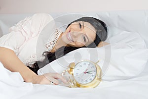 Beautiful brunette young woman blue eyes girl waking up lying in bed holding alarm clock happy smiling & looking at camera