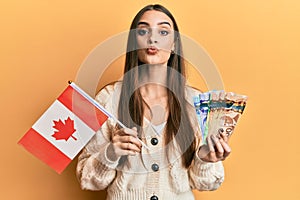 Beautiful brunette young woman holding canada flag and dollars looking at the camera blowing a kiss being lovely and sexy