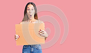 Beautiful brunette young woman holding cardboard banner with blank space scared and amazed with open mouth for surprise, disbelief