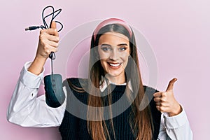 Beautiful brunette young woman holding computer mouse device smiling happy and positive, thumb up doing excellent and approval