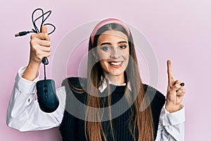 Beautiful brunette young woman holding computer mouse device smiling with an idea or question pointing finger with happy face,