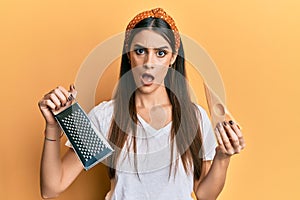 Beautiful brunette young woman holding grater and cheese in shock face, looking skeptical and sarcastic, surprised with open mouth