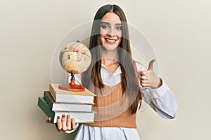 Beautiful brunette young woman holding pile of books and vintage world ball smiling happy and positive, thumb up doing excellent