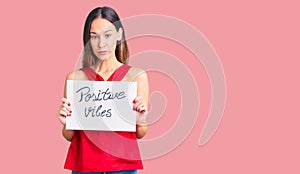 Beautiful brunette young woman holding positive vibes banner thinking attitude and sober expression looking self confident