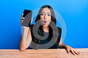 Beautiful brunette young woman holding smartphone showing blank screen scared and amazed with open mouth for surprise, disbelief