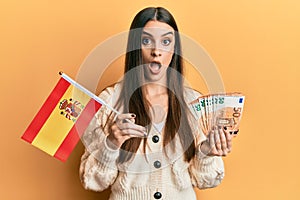 Beautiful brunette young woman holding spain flag and euros banknotes afraid and shocked with surprise and amazed expression, fear