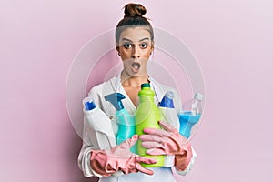 Beautiful brunette young woman wearing cleaner apron holding cleaning products afraid and shocked with surprise and amazed