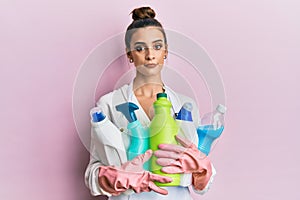 Beautiful brunette young woman wearing cleaner apron holding cleaning products relaxed with serious expression on face