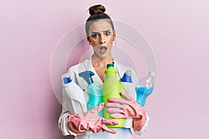 Beautiful brunette young woman wearing cleaner apron holding cleaning products in shock face, looking skeptical and sarcastic,