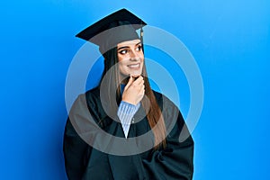 Beautiful brunette young woman wearing graduation cap and ceremony robe with hand on chin thinking about question, pensive