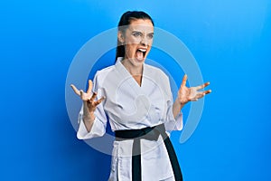 Beautiful brunette young woman wearing karate fighter uniform with black belt crazy and mad shouting and yelling with aggressive