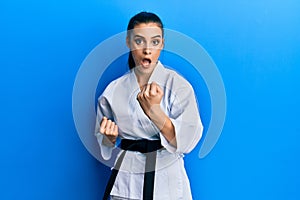 Beautiful brunette young woman wearing karate fighter uniform with black belt doing attack pose afraid and shocked with surprise