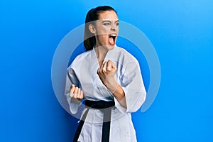 Beautiful brunette young woman wearing karate fighter uniform with black belt doing attack pose angry and mad screaming frustrated