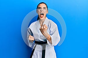 Beautiful brunette young woman wearing karate fighter uniform with black belt doing attack pose angry and mad screaming frustrated