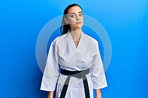 Beautiful brunette young woman wearing karate fighter uniform with black belt relaxed with serious expression on face