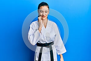 Beautiful brunette young woman wearing karate fighter uniform with black belt touching mouth with hand with painful expression