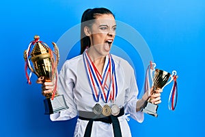 Beautiful brunette young woman wearing karate fighter uniform and medals holding trophy angry and mad screaming frustrated and