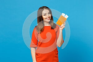 Beautiful brunette young woman wearing red orange dress hol in hand passport tickets isolated over trendy blue wall