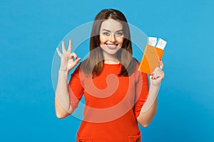 Beautiful brunette young woman wearing red orange dress hol in hand passport tickets isolated over trendy blue wall