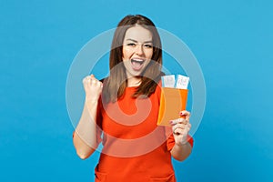 Beautiful brunette young woman wearing red orange dress hol in hand passport tickets isolated over trendy blue wall