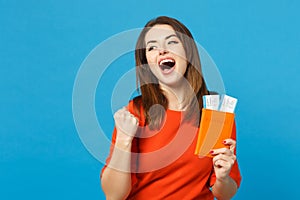 Beautiful brunette young woman wearing red orange dress hol in hand passport tickets isolated over trendy blue wall