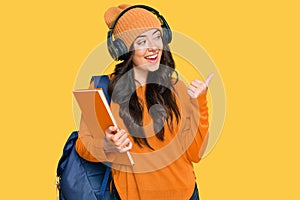 Beautiful brunette young woman wearing student backpack and holding books pointing thumb up to the side smiling happy with open