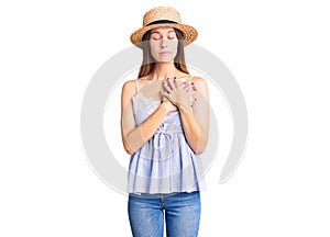 Beautiful brunette young woman wearing summer hat smiling with hands on chest with closed eyes and grateful gesture on face