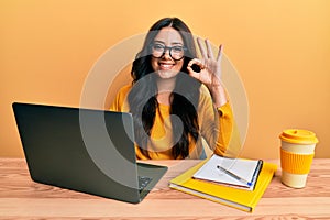 Beautiful brunette young woman working at the office wearing glasses doing ok sign with fingers, smiling friendly gesturing