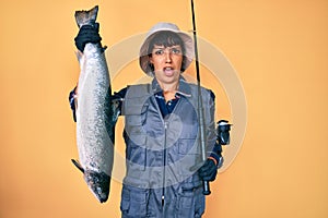 Beautiful brunettte fisher woman holding fishing rod and raw salmon clueless and confused expression