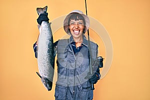 Beautiful brunettte fisher woman holding fishing rod and raw salmon sticking tongue out happy with funny expression