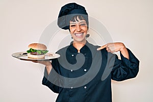 Beautiful brunettte woman chef holding burguer pointing finger to one self smiling happy and proud