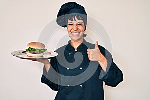 Beautiful brunettte woman chef holding burguer smiling happy and positive, thumb up doing excellent and approval sign