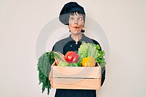 Beautiful brunettte woman chef holding fresh veggetables making fish face with mouth and squinting eyes, crazy and comical