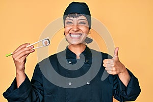 Beautiful brunettte woman chef holding sushi using chopsticks smiling happy and positive, thumb up doing excellent and approval