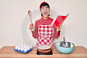 Beautiful brunettte woman cooking cake reading recipes book angry and mad screaming frustrated and furious, shouting with anger