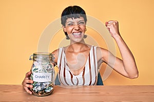 Beautiful brunettte woman holding charity jar with money screaming proud, celebrating victory and success very excited with raised