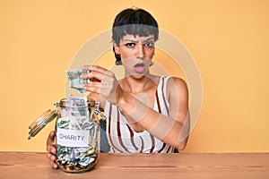 Beautiful brunettte woman holding charity jar with money in shock face, looking skeptical and sarcastic, surprised with open mouth