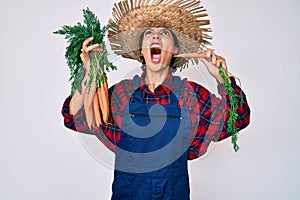 Beautiful brunettte woman wearing farmer clothes holding fresh carrots angry and mad screaming frustrated and furious, shouting