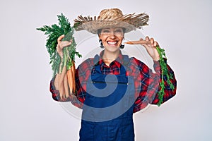 Beautiful brunettte woman wearing farmer clothes holding fresh carrots smiling with a happy and cool smile on face