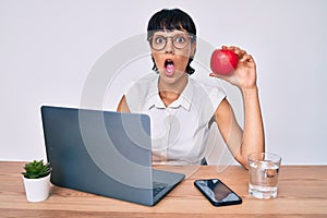 Beautiful brunettte woman working at the office eating healthy apple scared and amazed with open mouth for surprise, disbelief